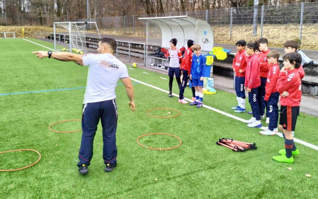 Coach Ray, Personal Trainer Konstanz, steht auf dem Sportplatz und macht mit jugendlichen Fussballern Geschwindigkeitstraining auf einem Kunstrasenplatz.
